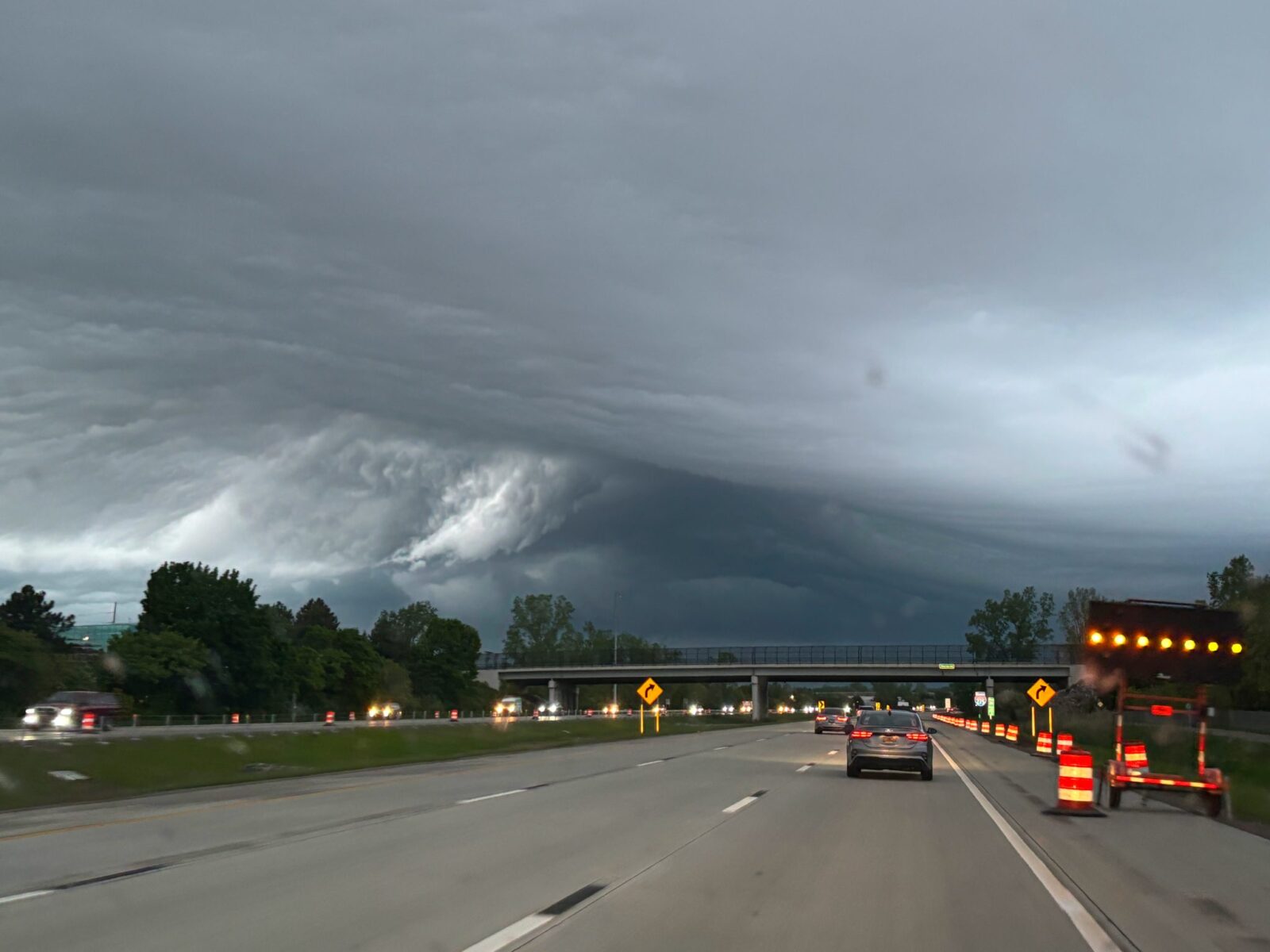 Ominous, dark storm clouds hang over a multi-lane highway with cars, a bridge, and construction signs